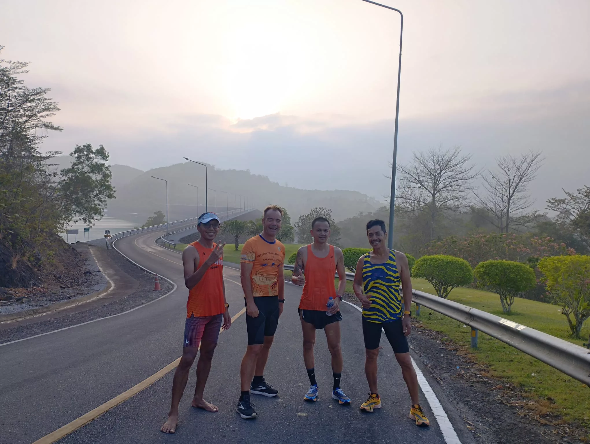 Eine Gruppe von Läufern joggt in Thailand auf einer gut asphaltierten Straße, im Hintergrund eine Berglandschaft mit See und aufgehender Sonne.