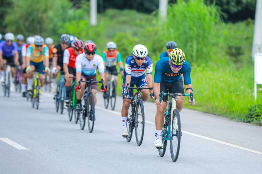 Eine größere Rennradgruppe fährt in Thailand auf einer sonnigen, gut asphaltierten Straße durch eine tropische Landschaft.